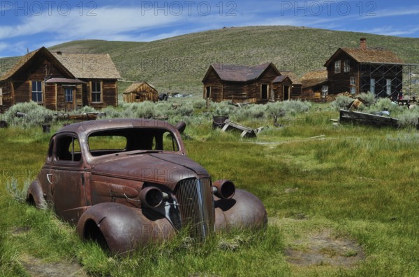 Rusty car wreck, Chevrolet Coupé, on green field in abandoned town with wooden houses, Bodie Ghost Town, California, USA