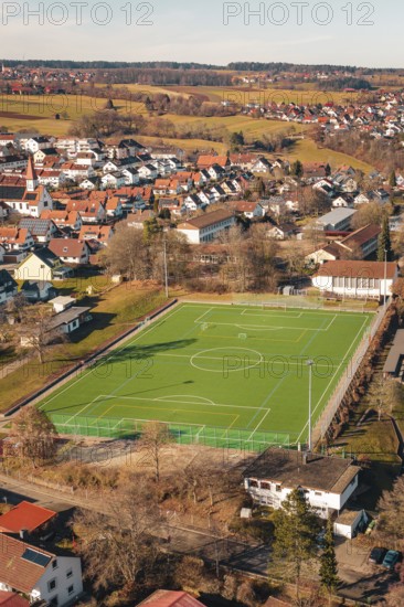 Green sports ground in a village surrounded by houses and hills seen from the air, Wimberg, Calw, Germany