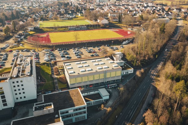 Aerial view of sports ground and school building with adjacent parking lot, nestled in an urban landscape in winter, Wimberg, Calw, Germany