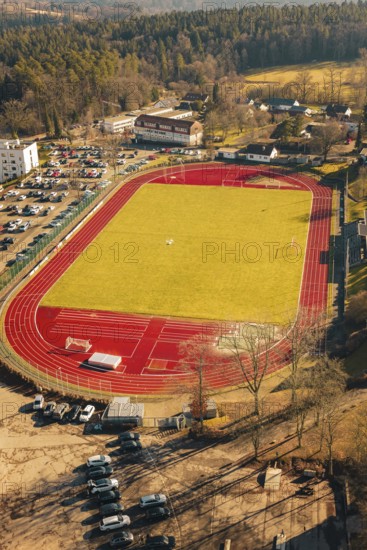 View of a large sports field with green field and red track, surrounded by cars and nature in winter, Wimberg, Calw, Germany