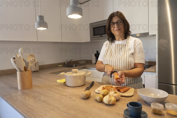 Smiling mature woman wearing apron peeling carrot while preparing vegetables in modern kitchen. Healthy homemade food, everyday lifestyle and cozy domestic environment