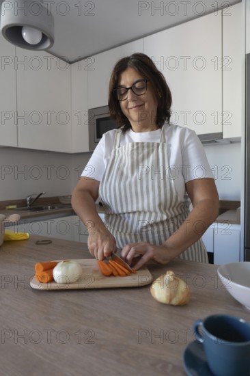 Mature woman wearing apron cutting carrots on wooden cutting board in modern kitchen. Healthy homemade food, everyday lifestyle and cozy domestic scene