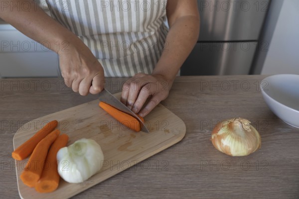Carrot being cut with a knife on a wooden cutting board while preparing vegetables in a home kitchen. Selective focus and natural light
