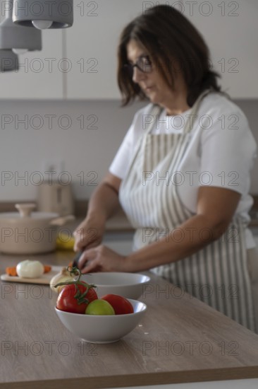 Selective focus on fresh tomatoes in foreground while woman chops onion in background. Home cooking and healthy food preparation concept