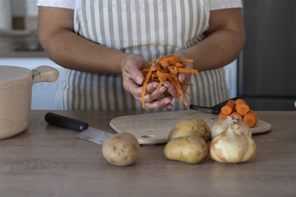 Close-up view of woman's hands holding carrot peels while peeling fresh carrots on a wooden board. Everyday cooking scene with natural light and selective focus