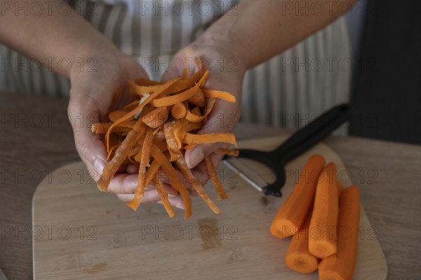 Close-up of hands holding fresh carrot peels while preparing vegetables on a wooden cutting board in a home kitchen. Natural light, selective focus, healthy cooking and zero waste food concept