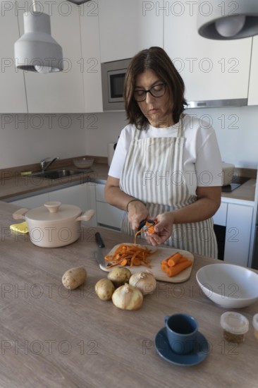 Woman wearing apron preparing vegetables for homemade soup while peeling carrots in modern kitchen. Simple ingredients and healthy home cooking concept