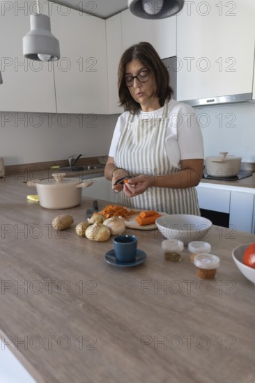 Cozy home kitchen scene with woman wearing apron peeling carrots and preparing vegetables. Comfort food and healthy lifestyle concept