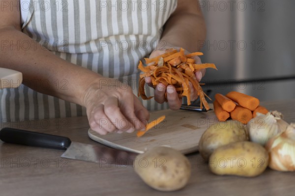 Hands holding freshly peeled carrot skins above a wooden cutting board with whole carrots and peeler. Natural light, shallow depth of field, home cooking concept