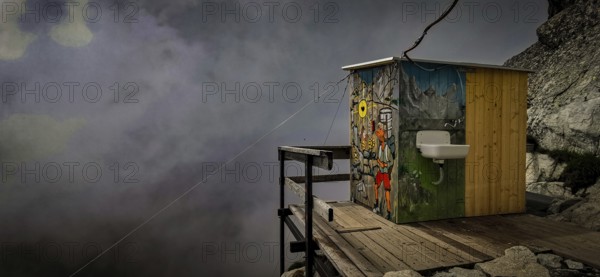 Colourful toilet of the Chata pod Rysmi mountain hut on a wooden platform in the middle of a mountainous, foggy landscape with wall art, graffiti, Rysy hiking, High Tatras, Slovakia