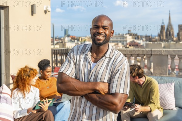 Happy young executive man smiling with arms crossed, standing outdoors on a coworking rooftop terrace with his diverse colleagues during a casual business meeting
