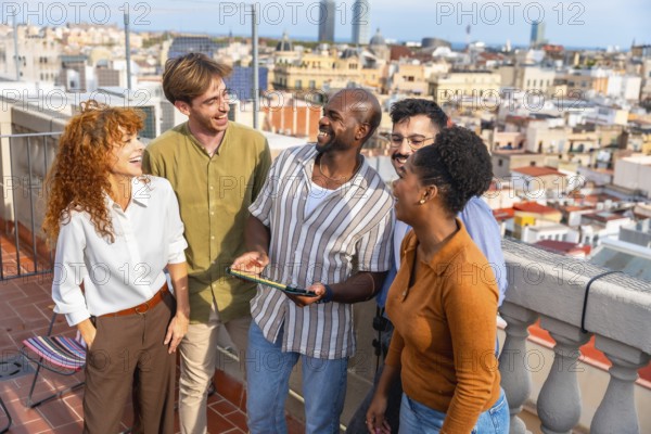 Diverse group of young professionals smiling and laughing on a barcelona rooftop, engaging in a casual work break or informal meeting while looking at a digital tablet