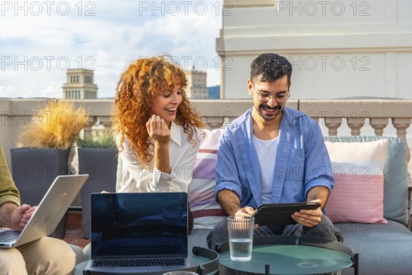 Young business colleagues working together on laptops and tablets, sharing ideas, and enjoying a casual outdoor meeting during their break on a modern urban rooftop coworking space