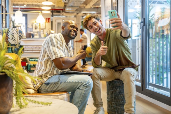 Diverse colleagues enjoying a casual break in a modern coworking space, smiling and giving thumbs up while one man takes a phone selfie, representing successful teamwork and collaboration