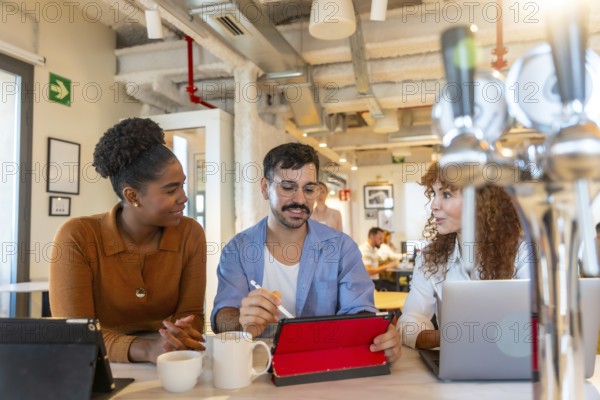 Diverse colleagues sitting at a high table, casually discussing a digital tablet and laptop during a break in a relaxed office or coworking environment