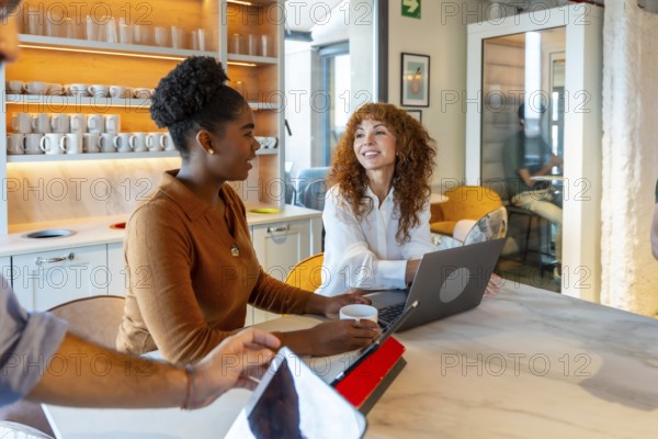 Multiracial businesswomen colleagues collaborating and discussing work around a laptop in a modern coworking kitchen while enjoying a coffee break, symbolizing teamwork and office life