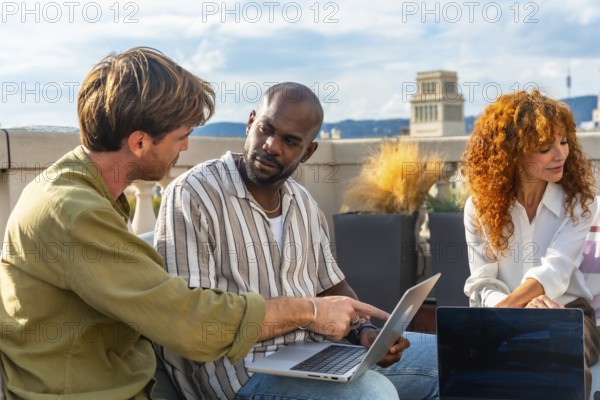 Diverse business professionals collaborating on a laptop during an informal rooftop meeting, sharing ideas and planning strategy in a sunny urban outdoor workspace - three people -