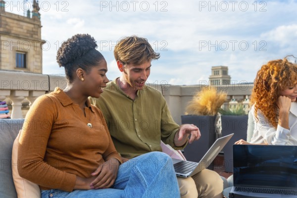 Diverse colleagues collaborate on a laptop during a rooftop meeting, sharing ideas and planning in a relaxed, sunny urban setting that blends work and social connection