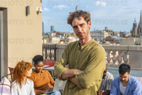 Confident man portraying leadership while standing with arms crossed, his diverse team having an informal coworking outdoor meeting on a city rooftop with buildings in the background