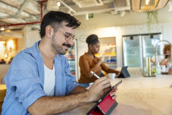 Man in casual attire smiling while using a digital tablet with a stylus to sketch ideas at a bar height table in a modern coworking space during a collaborative break