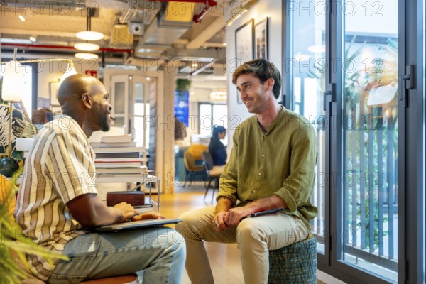 Two diverse male colleagues collaborate and chat during an informal break in a modern coworking office, sharing ideas, smiling and planning projects in a relaxed, professional setting