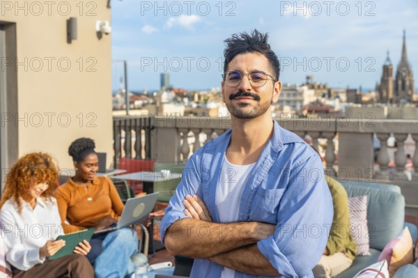 Young man with crossed arms and confident smile while diverse colleagues collaborate and chat during a sunny rooftop meeting in barcelona, casual modern teamwork and networking