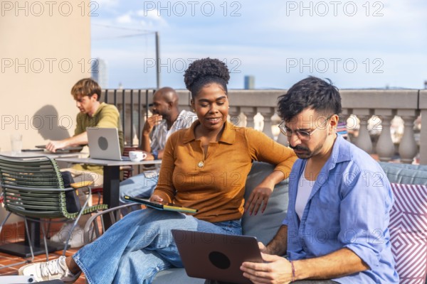 Diverse colleagues working and interacting on a rooftop terrace, using laptops and tablets while discussing projects in a modern outdoor office setting