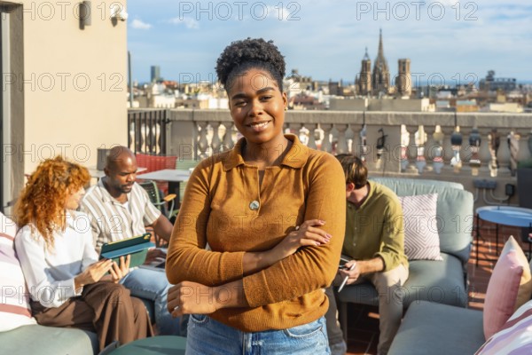Young black woman with an afro hairstyle smiles at the camera on a modern rooftop terrace while diverse colleagues work and interact in the blurred background, overlooking a city skyline