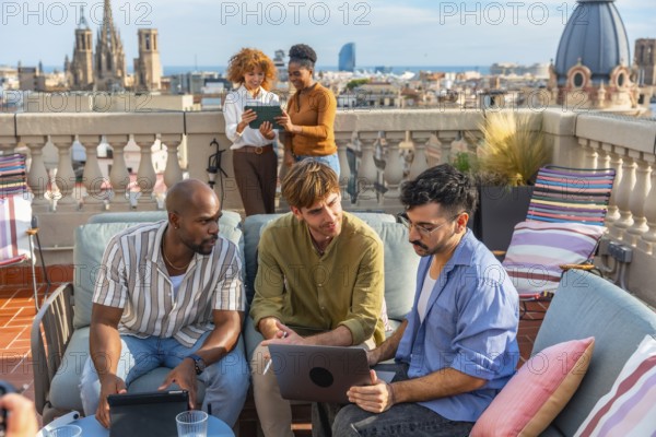 Diverse colleagues collaborate on a rooftop terrace in barcelona during a break, using laptop and tablet while discussing strategy and brainstorming against the city skyline