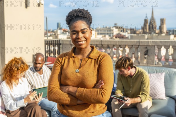 Confident black woman leader standing with arms crossed, smiling at the camera while her diverse business colleagues are having a relaxed outdoor discussion on a city rooftop