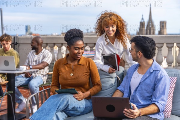 Diverse business professionals collaborating and networking on a barcelona rooftop terrace, using laptops and tablets for a casual outdoor meeting that blends teamwork and city lifestyle