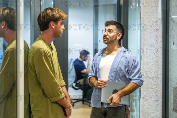 Two male colleagues engaging in an informal discussion during a break in a modern coworking space, with another person working in the background, representing teamwork and collaboration
