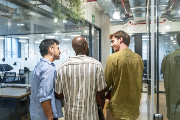 Three diverse men colleagues are engaging in conversation, networking, and discussing ideas while standing in a contemporary office or coworking space during a break