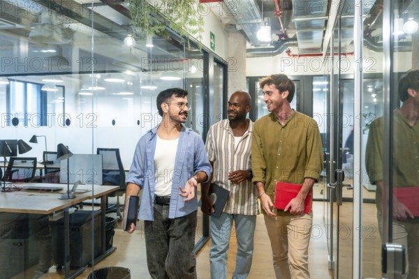 Three diverse male colleagues walking through a modern office corridor, smiling and engaging in conversation while carrying documents and tablets during a break