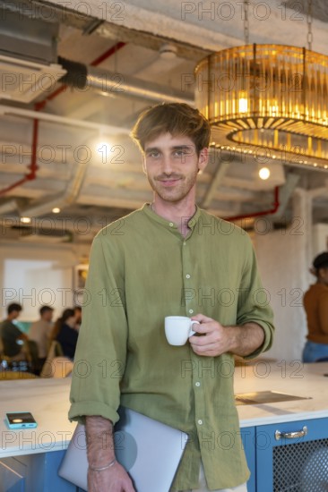 Young man standing in a modern coworking office, enjoying a coffee break while holding a laptop, surrounded by colleagues creating a dynamic work environment during a collaborative meeting