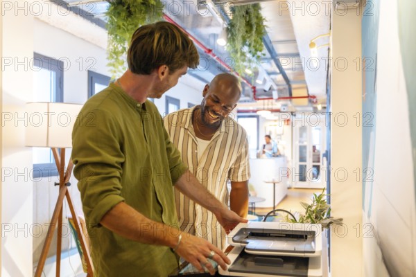 Male coworkers collaborating on a document at the printer, sharing a laugh and enjoying positive interaction in a modern coworking space, illustrating a friendly and productive work environment
