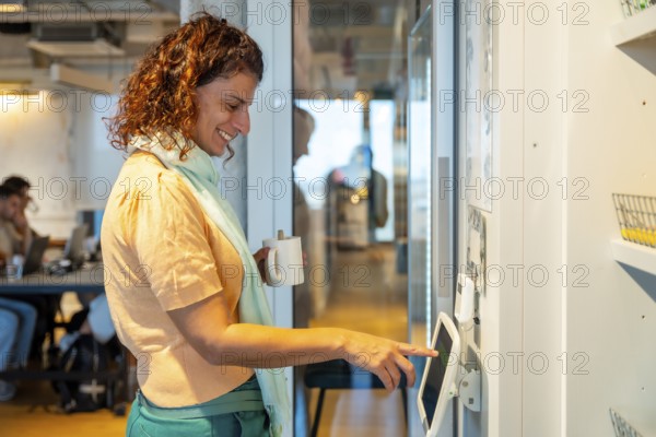 Woman standing in a modern office or coworking space, smiling and holding a coffee cup while using a digital tablet screen on a wall, showcasing a happy workflow and daily office life