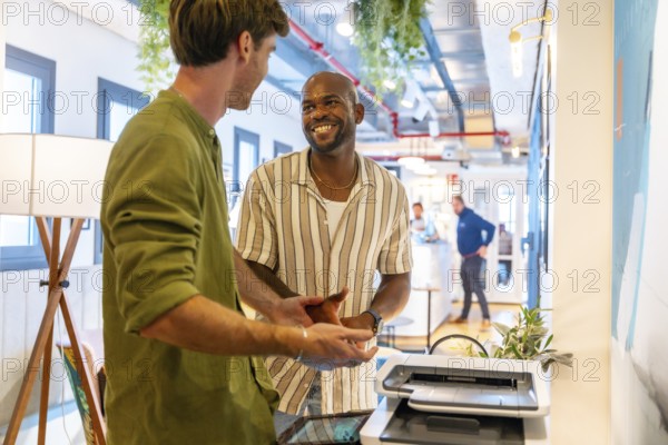 Two diverse male colleagues having a friendly discussion and smiling during a relaxed break in a modern, vibrant coworking office environment, fostering team collaboration and communication