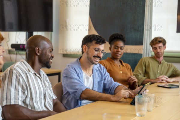 Diverse group of smiling colleagues interacting and working together at a conference table, using a tablet during a productive brainstorming session in a modern coworking space