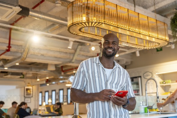 Smiling young black male professional resting in a modern coworking office space while checking his mobile phone, enjoying a relaxing tech enabled workplace break