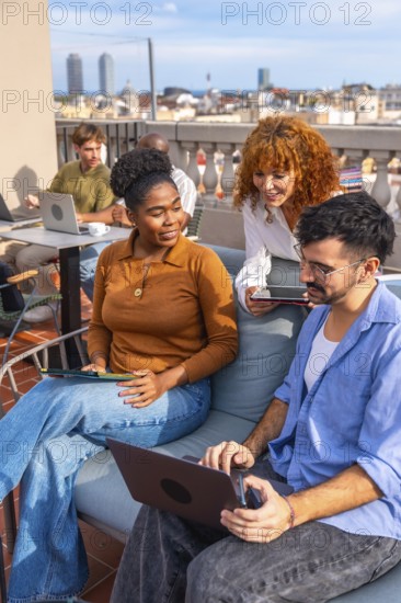 Diverse colleagues collaborating and sharing ideas while working on laptops and tablets during an informal meeting on an urban rooftop terrace with a city skyline