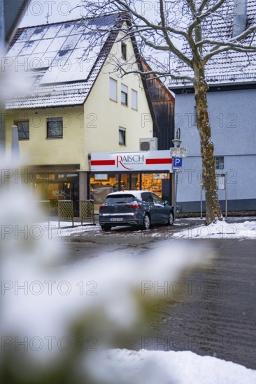 Snowy city street with parked car in front of a building with a shop, Gechingen, Calw district, Germany