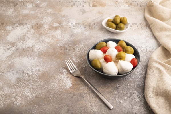 White Mozzarella cheese, with tomatoes and olives in blue bowl on brown concrete background and linen textile, side view, copy space