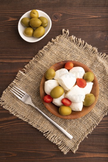 White Mozzarella cheese, with tomatoes and olives in wooden bowl on brown wooden background and linen textile, top view, flat lay, close up