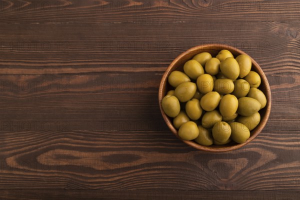 Fresh green olives in wooden bowl on brown wooden background. Top view, flat lay, copy space