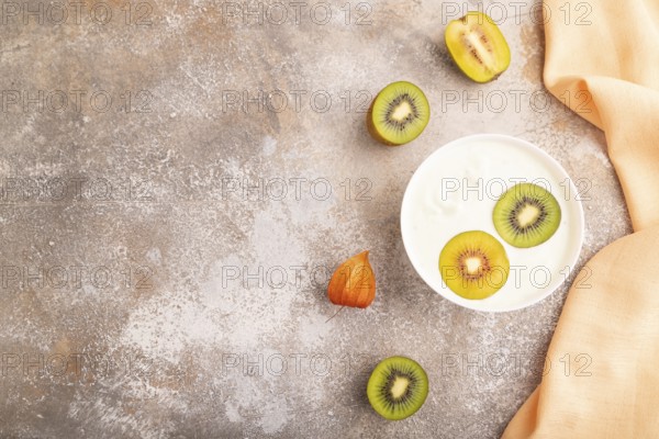 Yogurt with kiwi in white bowl on brown concrete background and orange linen textile, top view, flat lay, copy space, minimalism