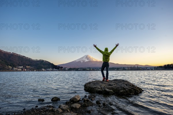 Young man stretching his arms in the air, standing on rocks in Lake Kawaguchi, view of Mount Fuji volcano at sunset, Yamanashi Prefecture, Japan