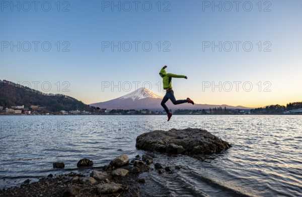 Young man jumping on rocks in Lake Kawaguchi, view of Mount Fuji volcano at sunset, Yamanashi Prefecture, Japan