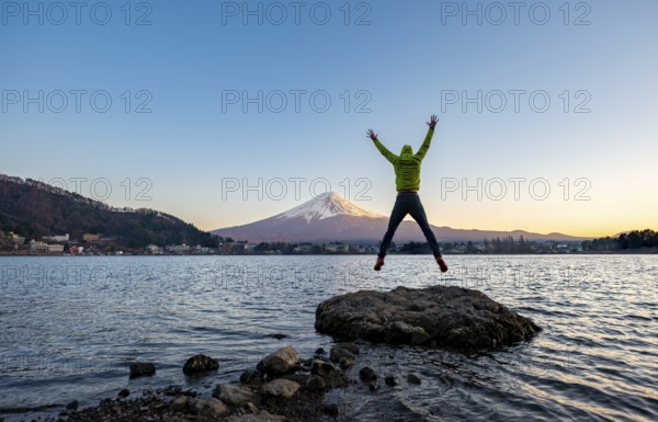 Young man jumping and stretching his arms in the air, standing on rocks in Lake Kawaguchi, view of Mount Fuji volcano at sunset, Yamanashi Prefecture, Japan