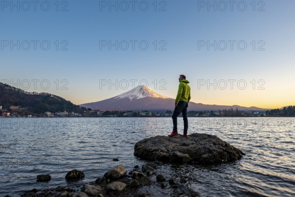Young man standing on rocks in Lake Kawaguchi, view of Mount Fuji volcano at sunset, Yamanashi Prefecture, Japan